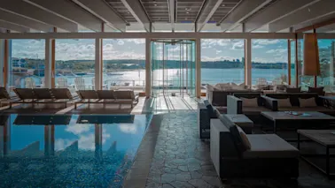 Innendørs bassengområde med solsenger og sitteplasser, med panoramautsikt over en solrik innsjø med båter og en fjern strand.