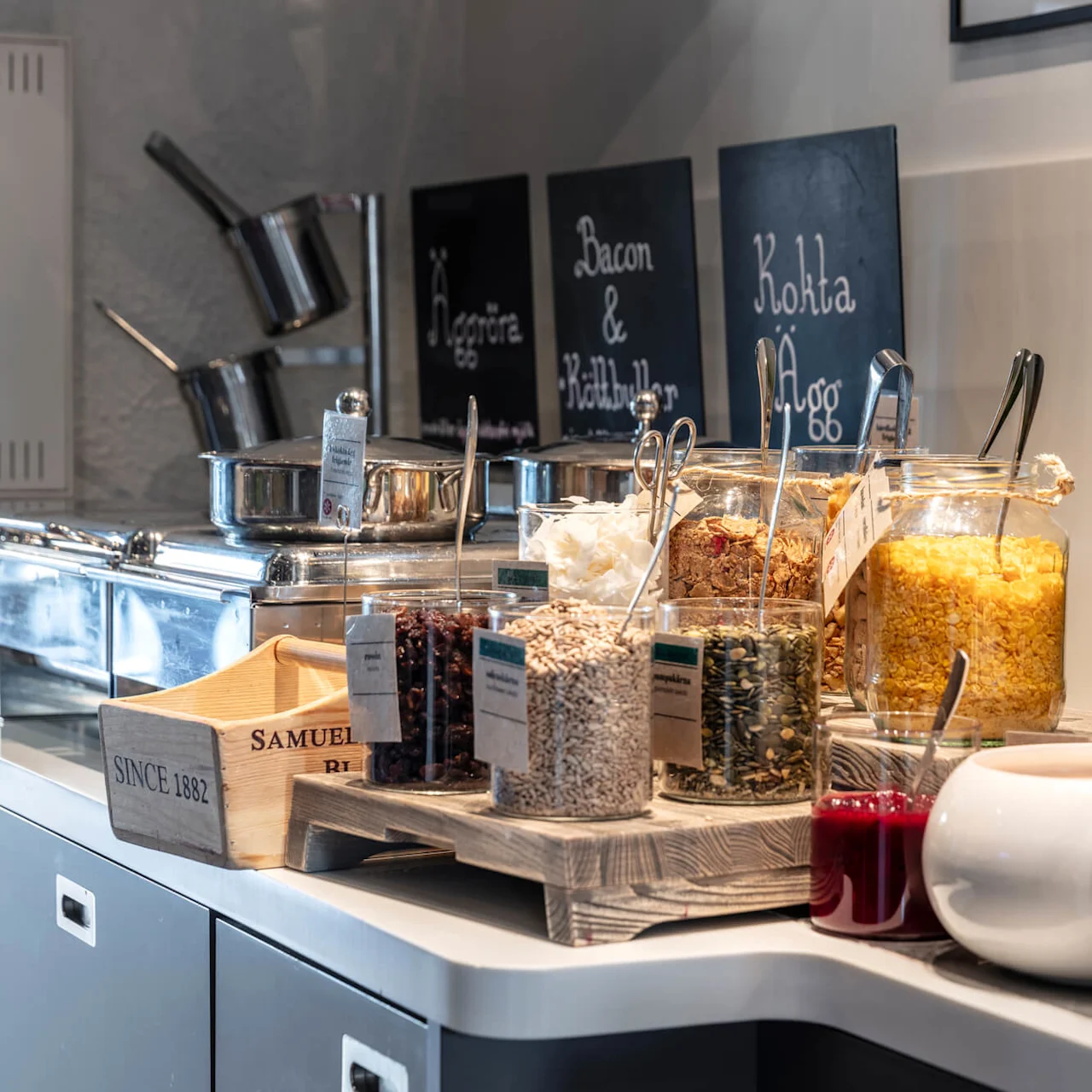 A breakfast buffet setup with cereals, sauces, and cooking pots, labeled with descriptions, in a cozy kitchen atmosphere. The boards in the background read "Bacon & Köttbullar" and "Kokta Ägg".