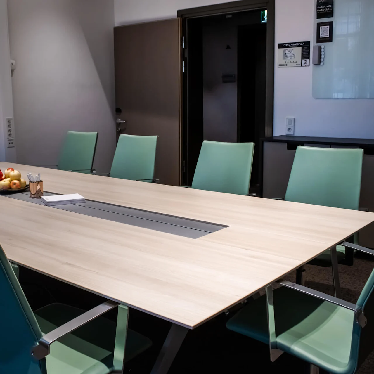 Et moderne konferanserom med et stort bord, turkise stoler og en svart skjerm på veggen.