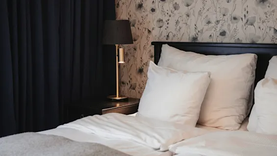 A cozy bedroom with a bed, nightstand, and lamp, featuring floral wallpaper and dark curtains for a restful atmosphere.