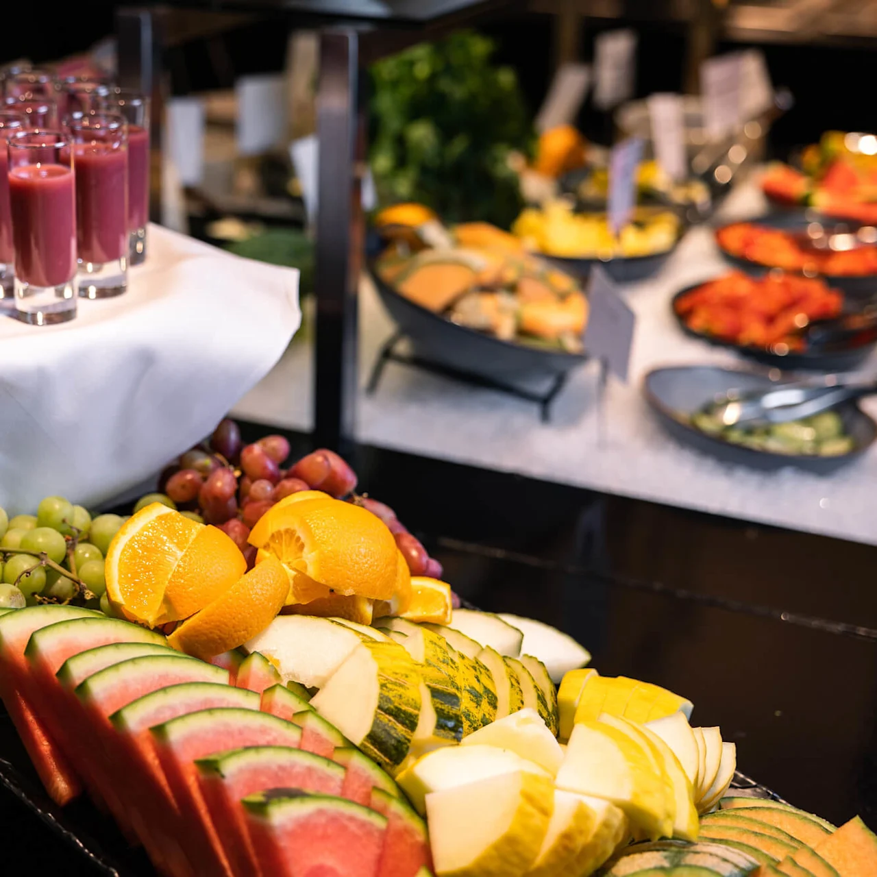 Glas fyldt med lilla smoothie står på en stofdækket bakke, med en farverig frugtplatte foran og flere retter i buffetbaggrunden.