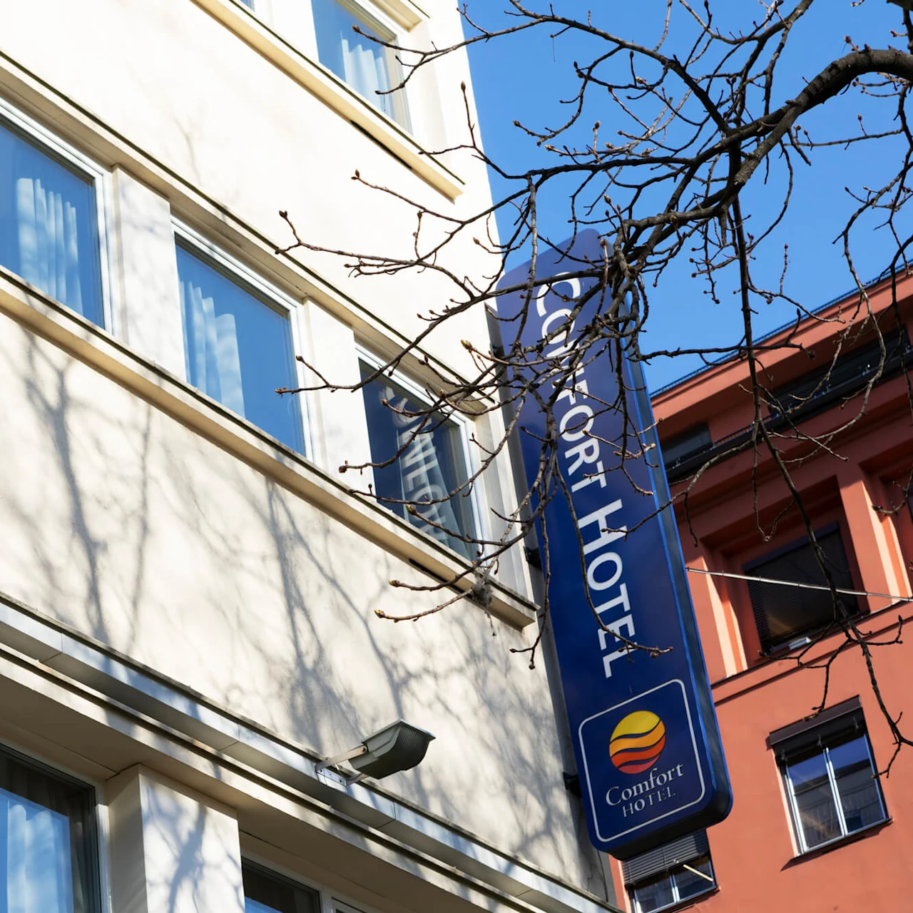 Hotellikyltti on kiinnitetty rakennukseen nimellä ja logolla, lehdettömän puun ja kirkkaansinisen taivaan välissä.