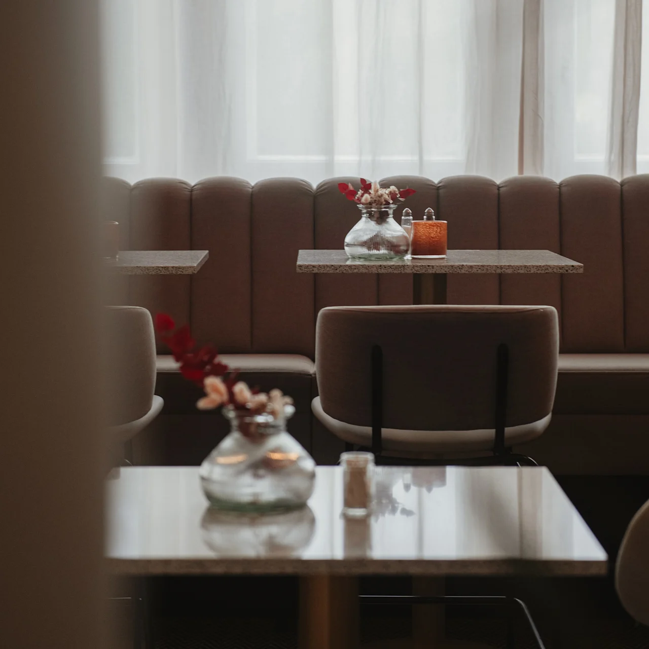 Tables hold glass vases with flowers, placed inside a softly lit café with upholstered seating and large curtains, creating a cozy and inviting atmosphere.