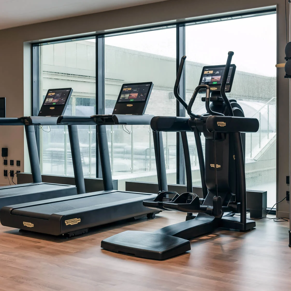 Løbebånd og en elliptisk maskine står stille i et moderne fitnesscenter med trægulve og store vinduer. "Technogym" mærke er synligt på udstyret.