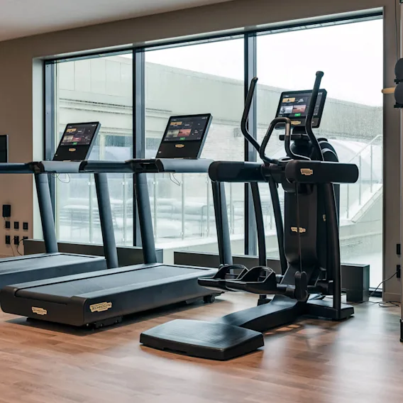 Treadmills and an elliptical machine stand idle in a modern gym with wooden floors and large windows. "Technogym" branding is visible on the equipment.