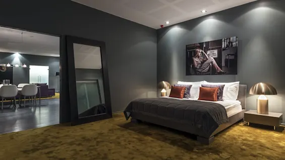 A modern bedroom features a neatly made bed with gray bedding, accented by brown pillows. A large mirror leans against a wall, while a stylish painting decorates above. 