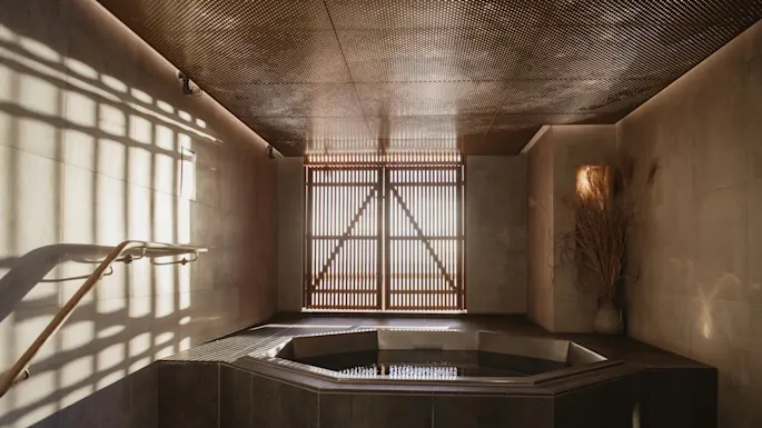 Et solfylt spa-rom med et sekskantet boblebad er nedsenket i gulvet, omgitt av steinfliser. En treskjerm sprer lys og kaster skygger på de beige veggene.