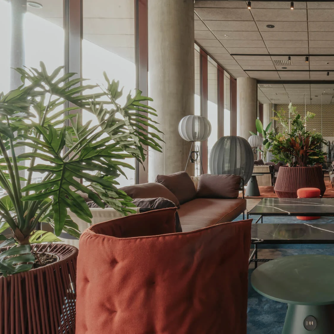 Bekväm sittgrupp med röda och bruna soffor, stora krukväxter och modern inredning i en rymlig, ljusfylld lobby med höga fönster och pelare.