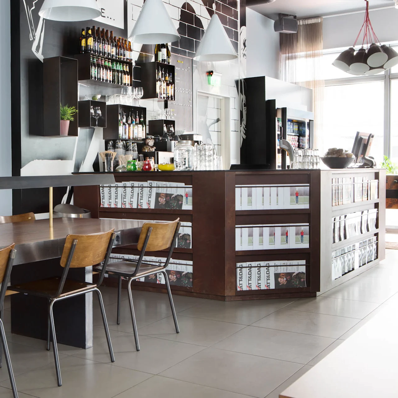 A large wooden table with chairs stands in front of a bar counter. Shelves filled with bottles and magazines are behind the counter. A television is mounted on the wall.