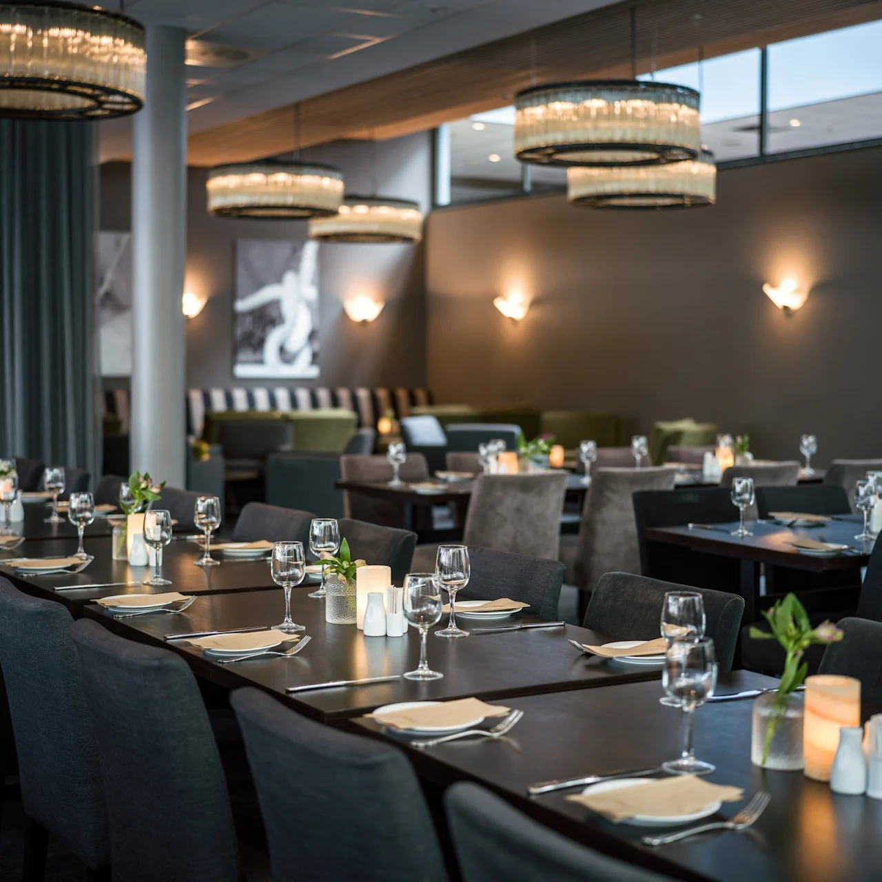 Matbord ordnade med prydligt placerade bordstabletter, glas och ljus. Taklampor lyser upp den moderna restaurangen med grå sittplatser och abstrakt konst på väggarna.