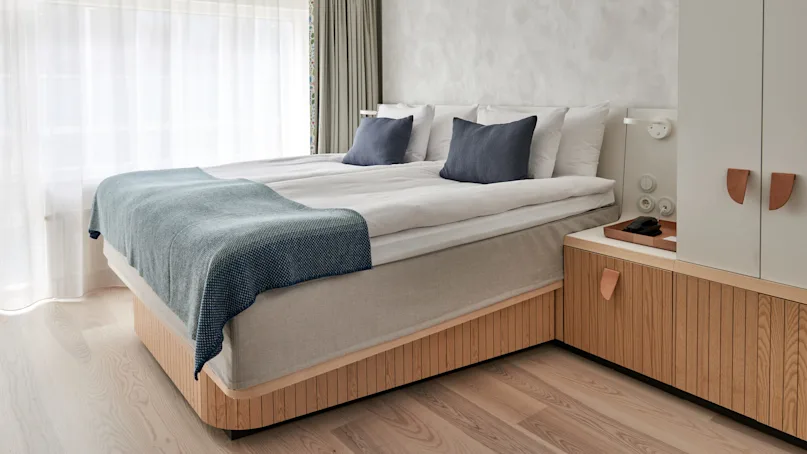 A neatly made bed with blue pillows and blanket rests in a bright, minimalist bedroom featuring light wood furniture and large, sheer-covered windows.