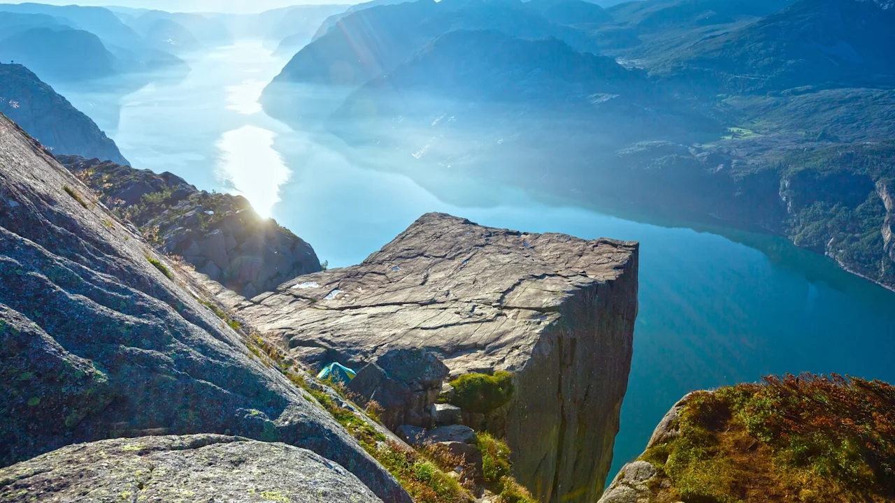 En steinete klippe stikker dramatisk ut over en fjord, med sterkt sollys som lyser opp den vidstrakte utsikten over fjerne fjell og vann nedenfor.