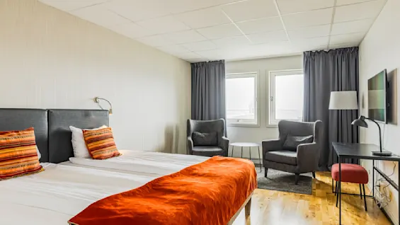 Et koselig hotellrom med dobbeltseng og livlige oransje detaljer, to lenestoler ved vinduet, og et lite skrivebord med en lampe. Lyset strømmer inn gjennom vinduene og skaper en innbydende atmosfære.