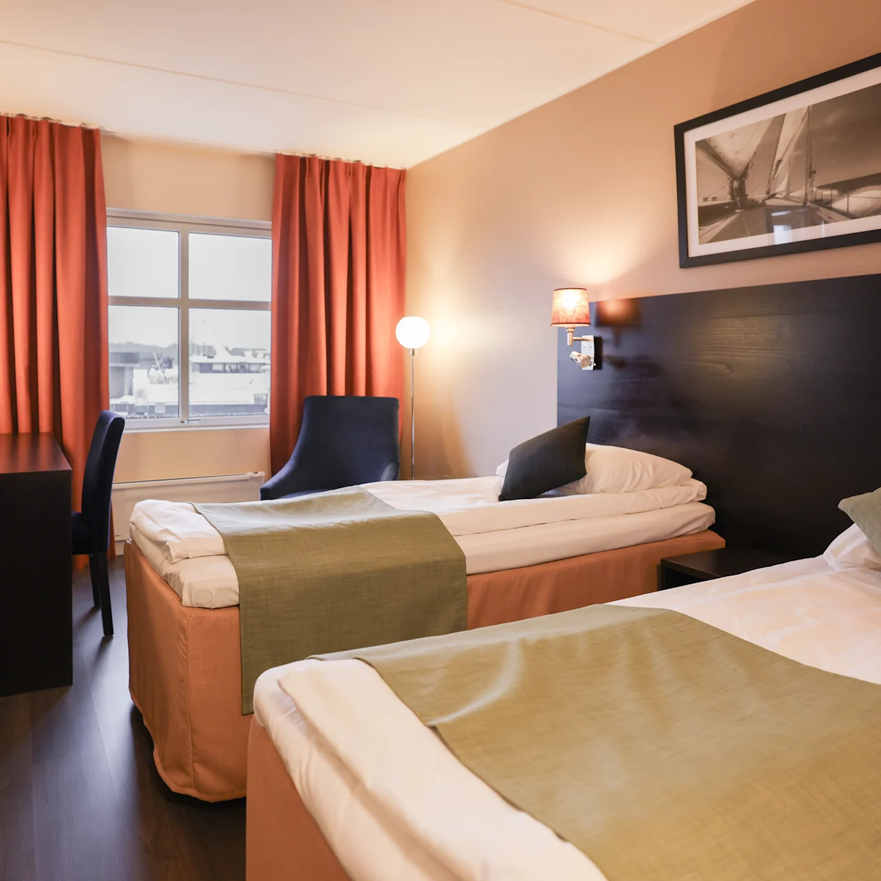 Two neatly made twin beds with green throws are positioned in a hotel room featuring a desk, chair, and a window with orange curtains. A black-and-white sailing photo adorns the wall.