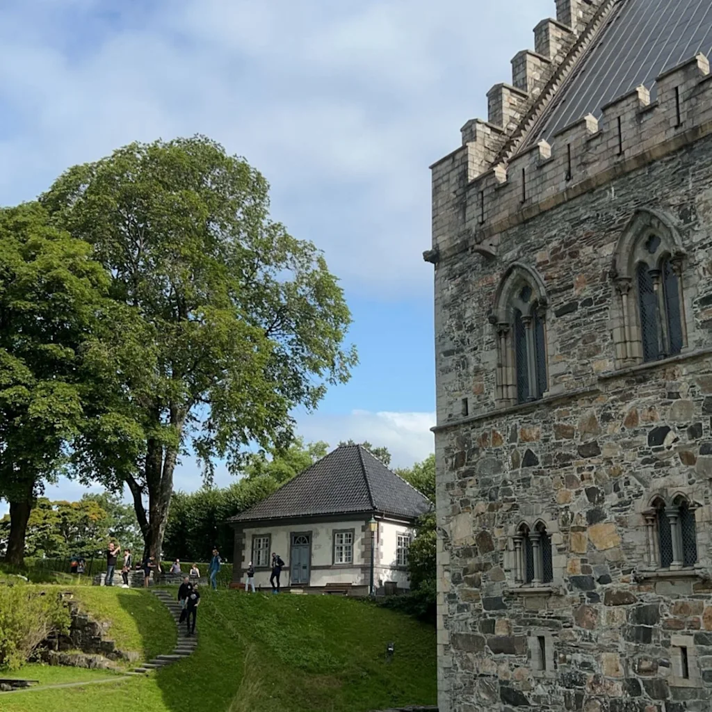 Bergenhus Festning: Historiallinen kivimuuri ja viehättävä valkoinen talo vihreällä kukkulalla, ihmisiä tutkimassa.