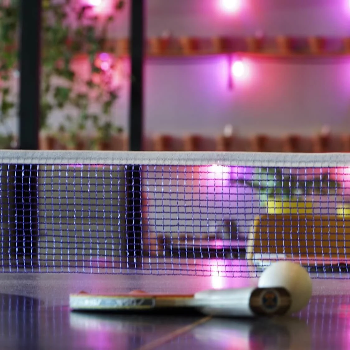 Ping-pong table with ball and paddle, dimly lit social ambiance.