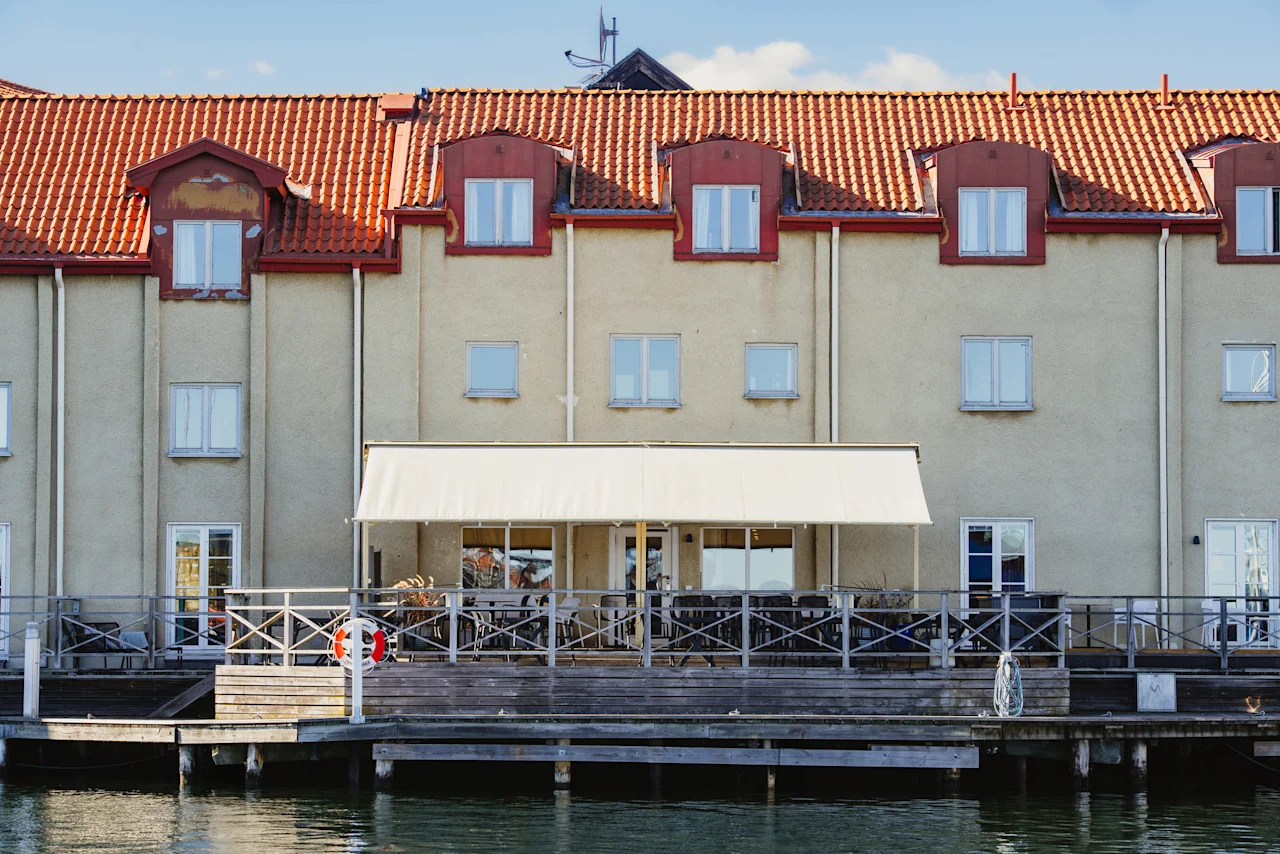 En bygning med rødt tegltag vender ud mod en vandterrasse med udendørs siddepladser og en hvid markise, hvilket antyder et behageligt spiseområde.