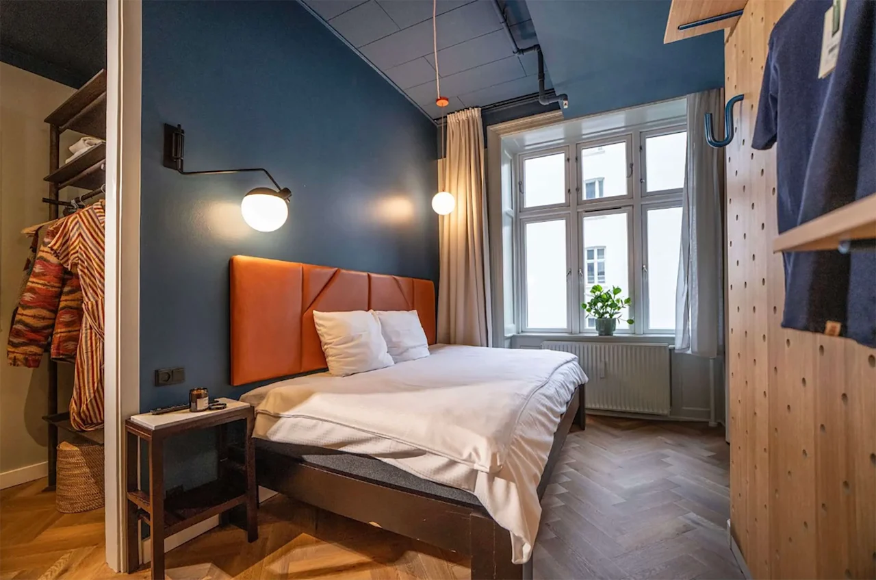 A modern hotel room with a double bed, blue walls, wooden floors, and a large window letting in natural light.
