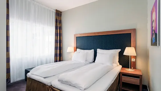 En seng med hvitt sengetøy er pent oppredd på et hotellrom. To nattbord med lamper står ved siden av sengen. Et vindu med lette gardiner og mønstrede gardiner slipper inn naturlig lys.