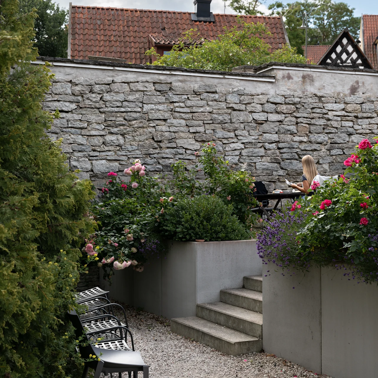 En kvinde sidder ved et bord og læser i en have med blomstrende blomster. Stentrapper og murer omgiver hende, og tagene er synlige under en delvist overskyet himmel.
