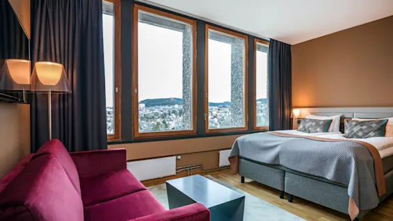A burgundy couch faces a large window with outdoor scenic views, beside a neatly made bed with gray bedding, in a cozy, modern room with wooden flooring and a soft rug.