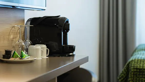 En svart kaffemaskin står på en bänkskiva med två glaskoppar, vita muggar och kaffepaket på en tallrik bredvid. En TV är monterad ovanför och gardiner är synliga.