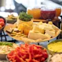 Ost- og grøntsagsfad arrangeret med skiver af ost, cherrytomater, rød peber og forskellige dyppelser. En olivenolieflaske og små glas med syltetøj ses i en lys spisestue.