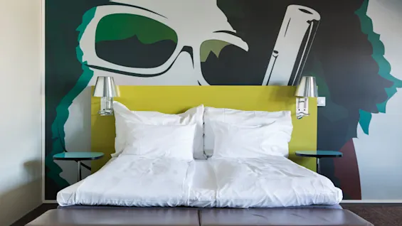 A neatly made double bed with white linens stands against a wall featuring a large, colorful mural of oversized sunglasses. Two side tables with metal lamps flank the bed.