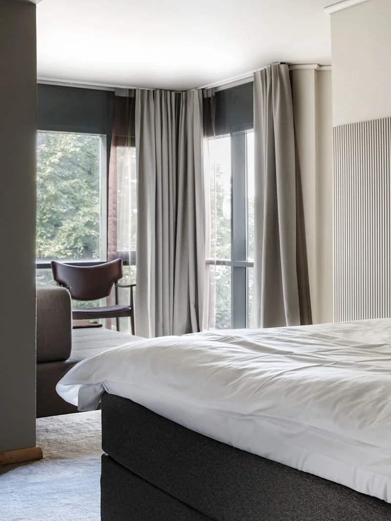 Siististi pedattu sänky valkoisilla vuodevaatteilla ja tyynyillä modernissa hotellihuoneessa, jossa on suuret ikkunat, harmaat verhot ja tuoli nurkassa lähellä sohvaa.