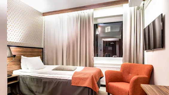 Ett mysigt hotellrum med en prydligt bäddad säng, fåtölj, gardiner och en väggmonterad TV bjuder på en bekväm tillfällig vistelse.