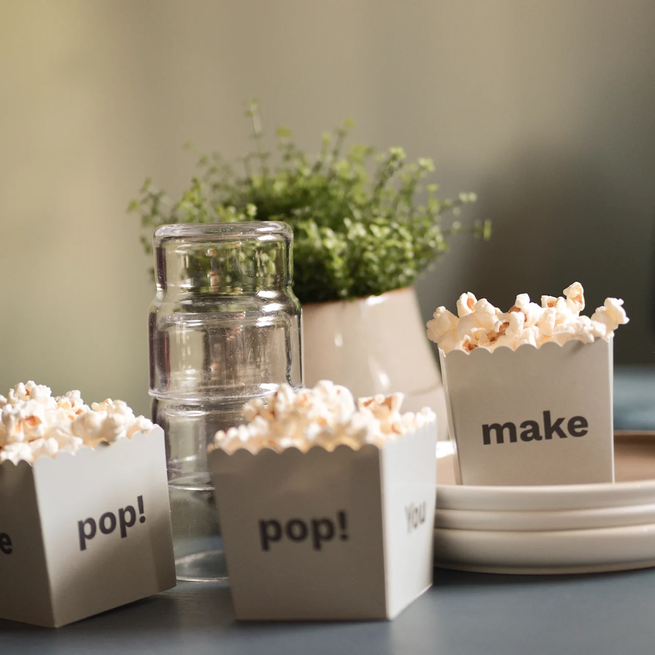 Popcornbeholdere mærket "me pop!", "pop!" og "make" står på et bord med tallerkener, glas og en lille potteplante i baggrunden.