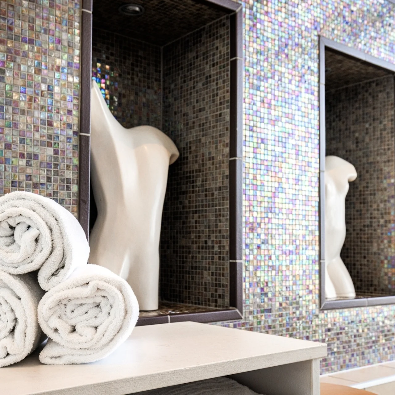 Three rolled white towels rest on a wooden surface. In the background, two white torso sculptures are displayed within mosaic-tiled niches. The setting appears to be a spa or relaxation area.
