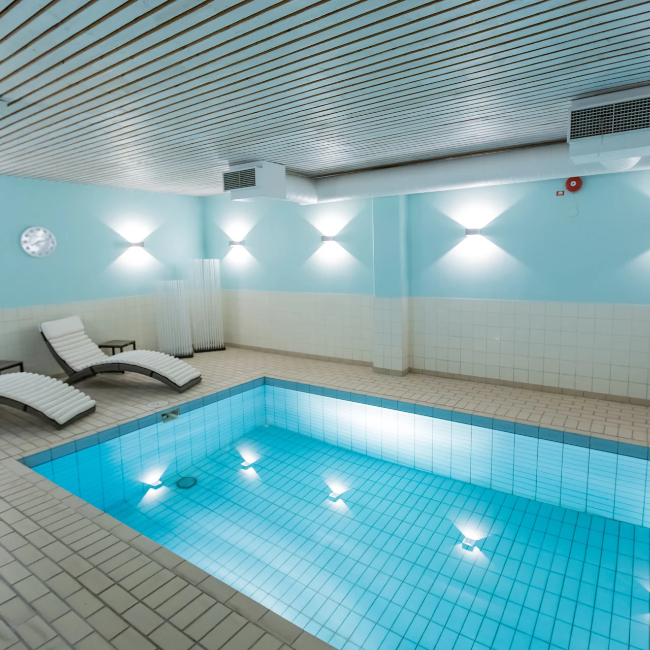 Indoor swimming pool is lit by submerged lights, reflecting on tiled walls. Two loungers are positioned nearby on a tiled floor, under soft overhead lighting.  