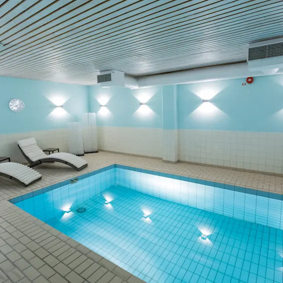 Inomhuspool lyses upp av underjordiska lampor och reflekteras på kaklade väggar. Två solstolar står i närheten på ett kaklat golv, under mjuk takbelysning.