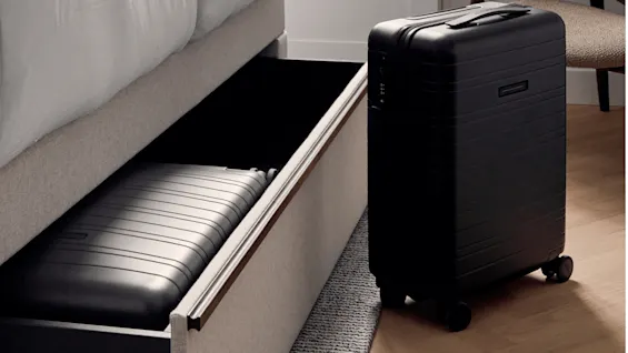 A black suitcase stands upright on a wooden floor next to an open drawer under a bed, containing another flat suitcase. A chair is visible in the background.