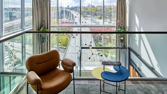 En brun læderstol vender mod en udsigt over en vej og bylandskab gennem store vinduer. Overhead er et farverigt graffitilofter i et moderne glasvægget rum med hængende lamper.
