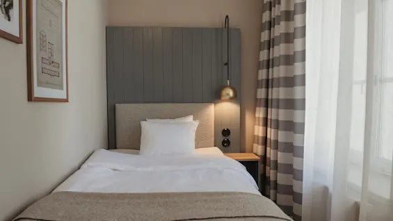 A cozy hotel room with a single bed, white bedding, and a warm throw. A golden lamp illuminates the bedside table.