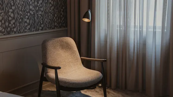 Viihtyisä nurkkaus, jossa on mukava nojatuoli, lattiavalaisin ja kuviollinen tapetti, ja pehmeä valo suodattuu läpikuultavien verhojen läpi.