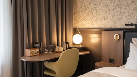 Ett skrivbord med en grön stol har en radio, glas och en sfärisk lampa, bredvid en säng. Mörka gardiner och ett mönstrat tapet skapar en mysig rumstämning.