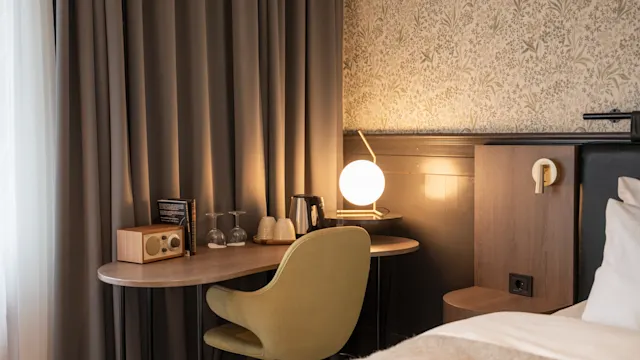 Et skrivebord med en grønn stol har en radio, glass og en sfærisk lampe, ved siden av en seng. Mørke gardiner og mønstret tapet skaper en koselig romatmosfære.