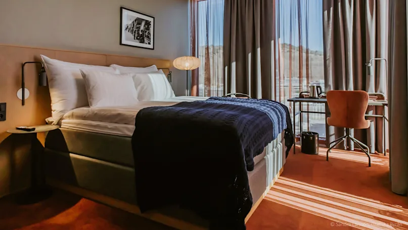 Bed with white pillows and blue blanket, situated by a window with curtains. Framed picture on wall; desk with chair, lamp, kettle nearby. Sunlight casts patterns on orange carpet.
