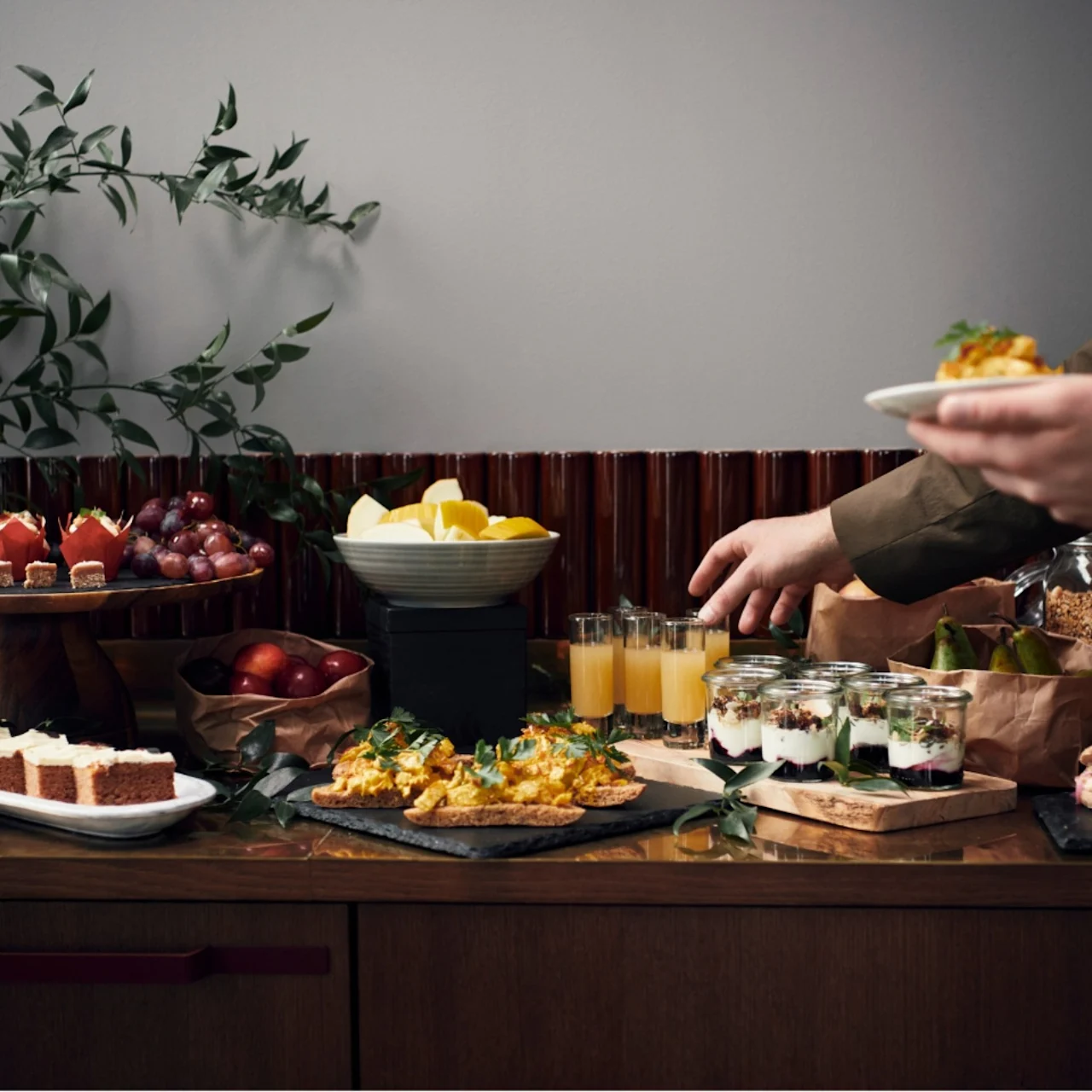 En person i en kostym tar ett glas apelsinjuice från ett träbord fyllt med olika sorters mat och desserter, dekorerat med blommor och grönt.