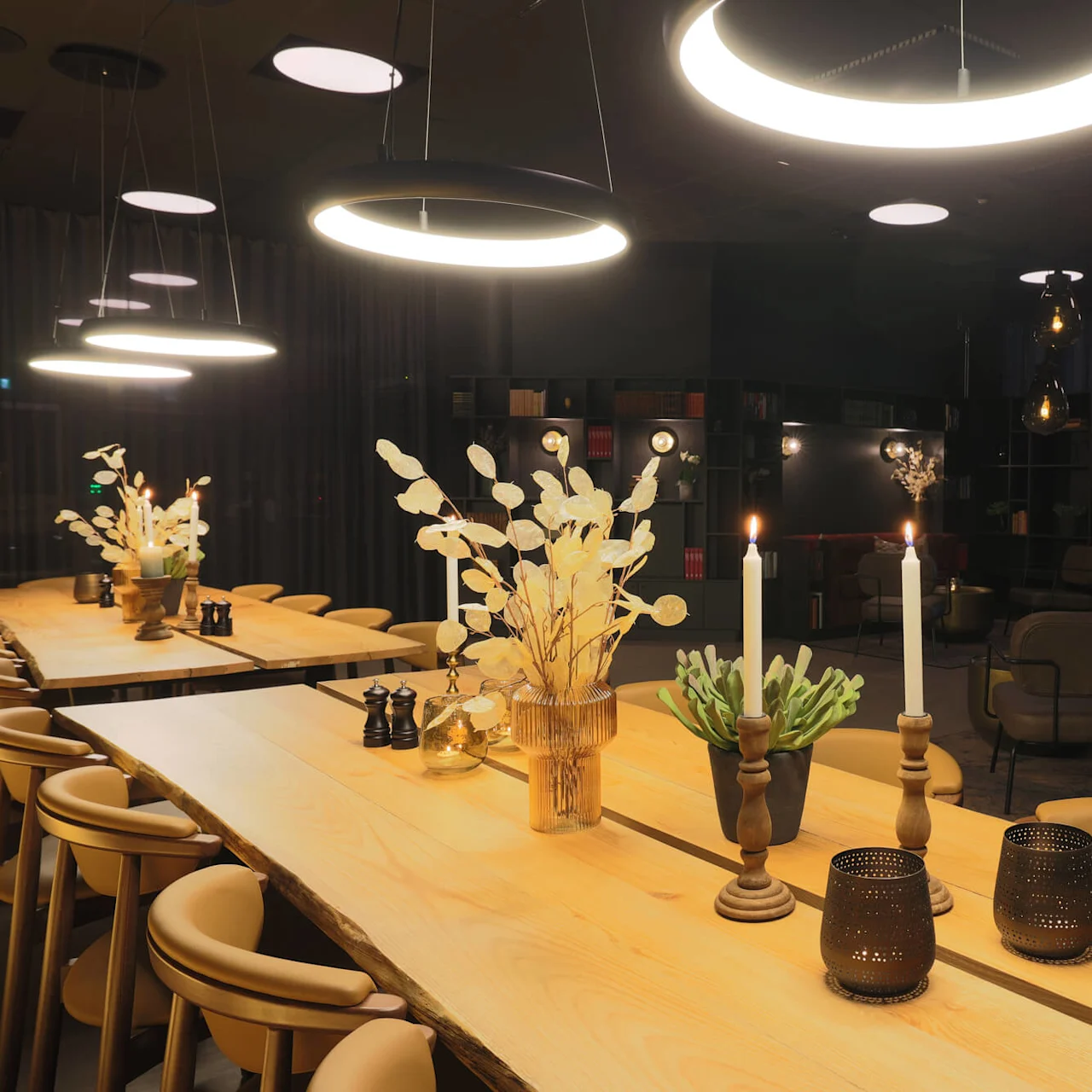 A well-lit modern interior with circular hanging lamps above wooden tables adorned with candles and plants, suggesting a cozy and stylish ambience.