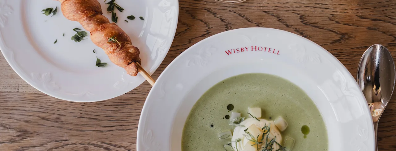 Grøn suppe pyntet med fløde og krydderurter i en tallerken mærket "WISBY HOTELL," ved siden af et spyd på en lille tallerken, altsammen på et træbord med et glas hvidvin.