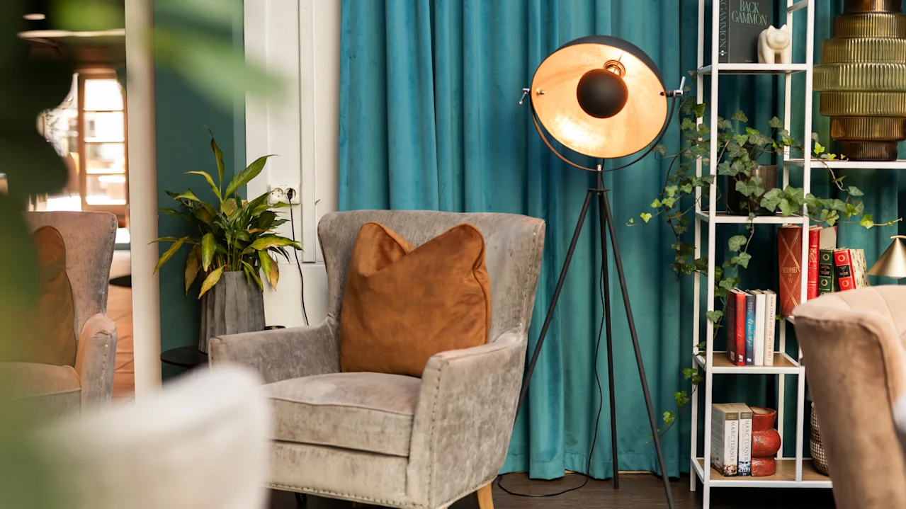 Lænestol med brun pude står i rum med blå gardiner. Lampe på stativ står ved siden af. Reol med bøger og dekorative genstande står i hjørnet. Frodig plante komplementerer det hyggelige opstilling.
