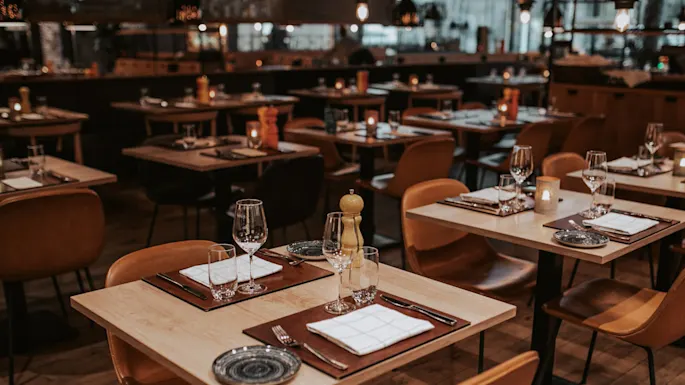 Bord dekket for middag, med vinglass, tallerkener og brettede servietter, skaper en koselig atmosfære i en varmt belyst restaurant med tregulv og pent arrangerte stoler.