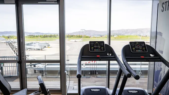 Treadmills positioned in front of large windows overlooking an airport runway with parked planes. Bright daylight illuminates the fitness area. No text visible.