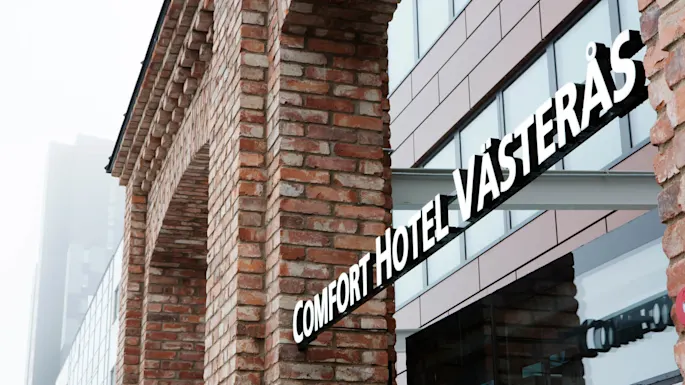 Teglstensbygningshjørne med skilt som leser "COMFORT HOTEL VÄSTERÅS" i et urbant miljø.