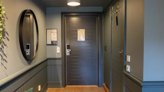 En mørktrædør i et hotelværelse, udstyret med en elektronisk lås, står lukket mellem to panelvægge og en loftlampe. Et rundt spejl og små meddelelser udsmykker væggene.