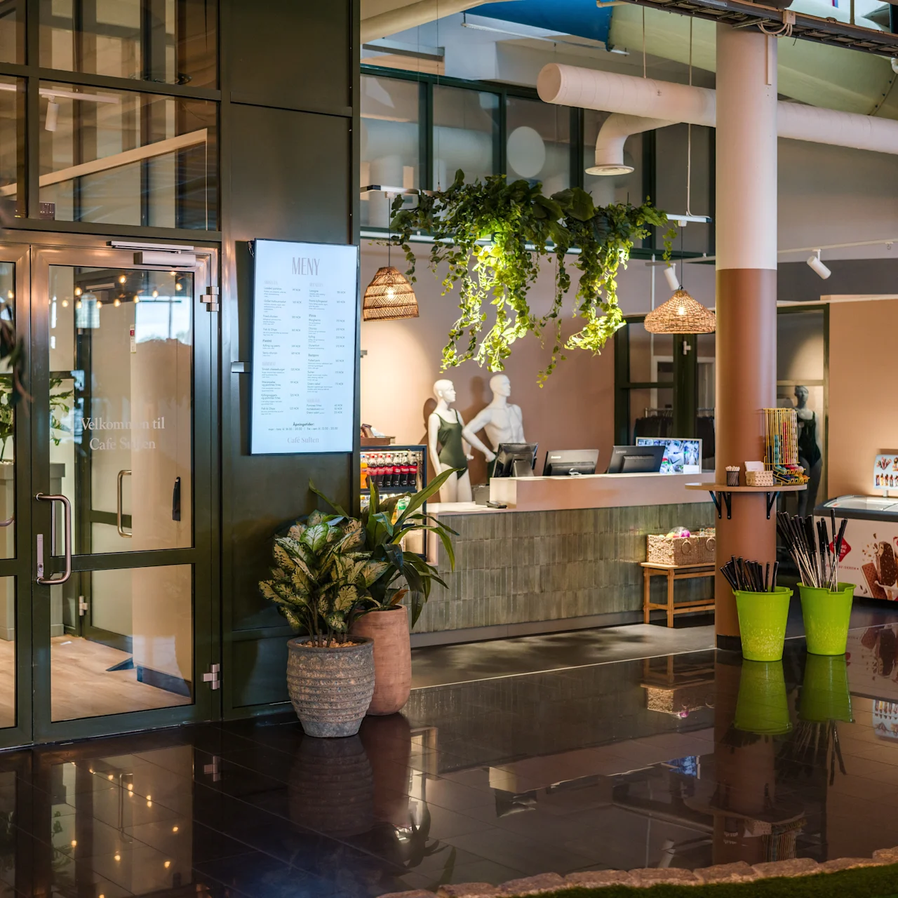 Reception desk with two mannequins behind it, under hanging plants and light fixtures. Adjacent glass door leads to another room. Potted plants and a display stand in the modern interior.